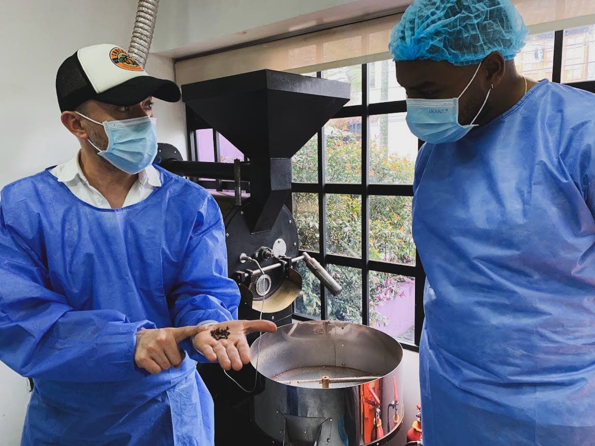 Dos visitantes con bata y tapabocas observan granos de cacao tostado junto al tostador de la planta