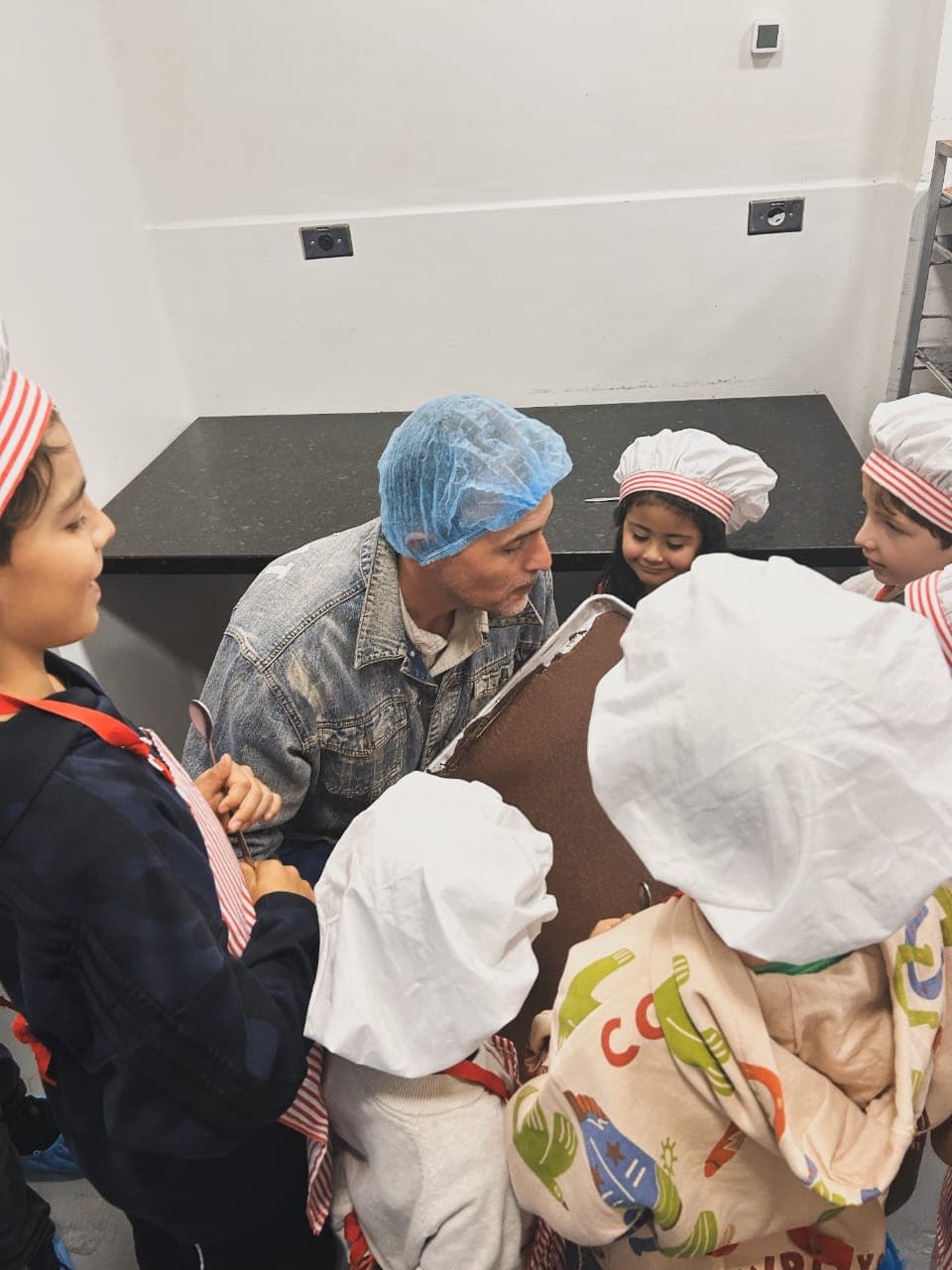 Niños con gorro de chef y delantal rodean al chocolatero que les muestra una bandeja de chocolate